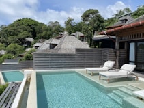 A serene outdoor setting featuring a pool with clear water and two white lounge chairs. The area is surrounded by lush greenery and modern wooden structures with sloped roofs. The sky is partly cloudy, adding to the peaceful ambiance.