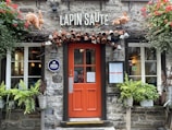 A charming exterior of a restaurant with a rustic stone wall and a bright orange door. Above the door, there are decorative hanging mugs and two sculpted rabbits. Plants and flowers adorn the entrance, adding to the cozy ambiance.