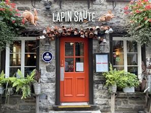 A charming exterior of a restaurant with a rustic stone wall and a bright orange door. Above the door, there are decorative hanging mugs and two sculpted rabbits. Plants and flowers adorn the entrance, adding to the cozy ambiance.