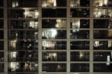 A multi-story residential building with large glass windows revealing the interior of various apartments. Most of the rooms are illuminated with warm indoor lighting, some darker than others. The windows display a variety of furniture and decorative items, highlighting different living spaces. There are balconies attached to a few apartments, adding to the urban architectural aesthetic.