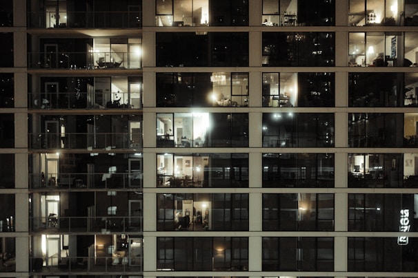 A multi-story residential building with large glass windows revealing the interior of various apartments. Most of the rooms are illuminated with warm indoor lighting, some darker than others. The windows display a variety of furniture and decorative items, highlighting different living spaces. There are balconies attached to a few apartments, adding to the urban architectural aesthetic.