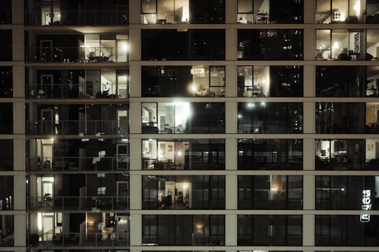 A multi-story residential building with large glass windows revealing the interior of various apartments. Most of the rooms are illuminated with warm indoor lighting, some darker than others. The windows display a variety of furniture and decorative items, highlighting different living spaces. There are balconies attached to a few apartments, adding to the urban architectural aesthetic.