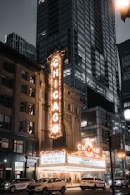 A bustling urban scene at night with a brightly lit theater marquee displaying the word 'CHICAGO' in large letters. The theater is situated among tall buildings in a city environment, with cars and pedestrians visible on the street. The marquee lists upcoming performances by various artists.
