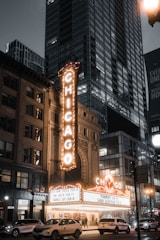 A bustling urban scene at night with a brightly lit theater marquee displaying the word 'CHICAGO' in large letters. The theater is situated among tall buildings in a city environment, with cars and pedestrians visible on the street. The marquee lists upcoming performances by various artists.
