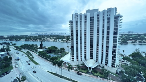 A tall, modern high-rise building stands prominently near a waterfront, surrounded by lush greenery and palm trees. The building is flanked by a wide road with several cars, and stretches of water and distant skyscrapers are visible in the background under a cloudy sky.