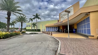 An outdoor view of a shopping center with prominent palm trees lining the pathway. The complex has a large sign for Old Navy Outlet and Dolphin Mall, with modern architectural design elements and a cloudy sky overhead.