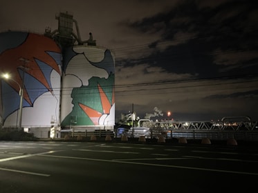 A large industrial structure, possibly a storage tank or silo, is painted with vibrant, abstract shapes in red, blue, white, and green hues. The setting appears to be at night, with surrounding infrastructure such as power lines, poles, and industrial plumbing visible. The sky is mostly dark with some illuminated clouds and distant lights from industrial activity.