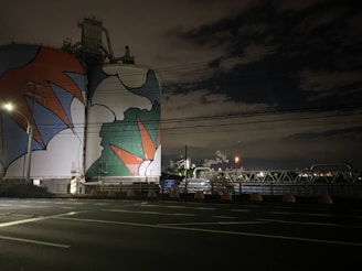 A large industrial structure, possibly a storage tank or silo, is painted with vibrant, abstract shapes in red, blue, white, and green hues. The setting appears to be at night, with surrounding infrastructure such as power lines, poles, and industrial plumbing visible. The sky is mostly dark with some illuminated clouds and distant lights from industrial activity.