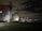 A large industrial structure, possibly a storage tank or silo, is painted with vibrant, abstract shapes in red, blue, white, and green hues. The setting appears to be at night, with surrounding infrastructure such as power lines, poles, and industrial plumbing visible. The sky is mostly dark with some illuminated clouds and distant lights from industrial activity.
