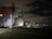 A large industrial structure, possibly a storage tank or silo, is painted with vibrant, abstract shapes in red, blue, white, and green hues. The setting appears to be at night, with surrounding infrastructure such as power lines, poles, and industrial plumbing visible. The sky is mostly dark with some illuminated clouds and distant lights from industrial activity.