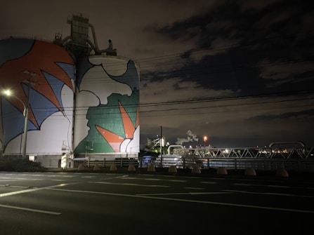 A large industrial structure, possibly a storage tank or silo, is painted with vibrant, abstract shapes in red, blue, white, and green hues. The setting appears to be at night, with surrounding infrastructure such as power lines, poles, and industrial plumbing visible. The sky is mostly dark with some illuminated clouds and distant lights from industrial activity.