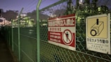 A chain-link fence topped with barbed wire runs alongside a walkway. Attached to the fence are warning signs in multiple languages, signaling restricted access and the presence of security cameras. Industrial structures are visible behind the fence, illuminated by artificial lighting against a darkening evening sky.