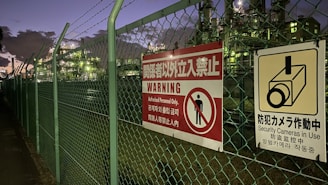 A chain-link fence topped with barbed wire runs alongside a walkway. Attached to the fence are warning signs in multiple languages, signaling restricted access and the presence of security cameras. Industrial structures are visible behind the fence, illuminated by artificial lighting against a darkening evening sky.