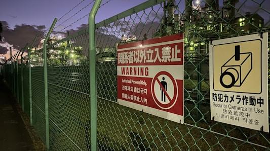 A chain-link fence topped with barbed wire runs alongside a walkway. Attached to the fence are warning signs in multiple languages, signaling restricted access and the presence of security cameras. Industrial structures are visible behind the fence, illuminated by artificial lighting against a darkening evening sky.