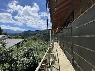 A partially constructed building with scaffolding on its side, set against a background of lush green hills and a clear blue sky with scattered clouds. The foreground shows construction materials and tools, while the surrounding area is filled with dense foliage and a nearby building with a metal roof.