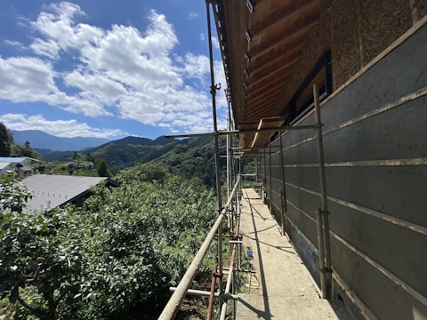 A partially constructed building with scaffolding on its side, set against a background of lush green hills and a clear blue sky with scattered clouds. The foreground shows construction materials and tools, while the surrounding area is filled with dense foliage and a nearby building with a metal roof.