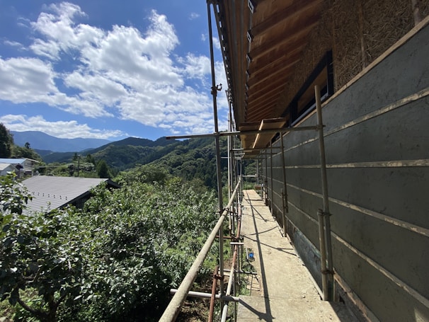 A partially constructed building with scaffolding on its side, set against a background of lush green hills and a clear blue sky with scattered clouds. The foreground shows construction materials and tools, while the surrounding area is filled with dense foliage and a nearby building with a metal roof.