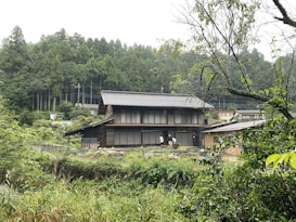 A traditional Japanese-style house situated in a lush, green forested area. The house features a dark tiled roof and large wooden sliding doors. Several trees and shrubs surround the building, adding to the serene, natural atmosphere.