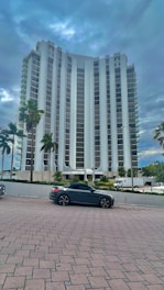 A tall, modern high-rise building with a sleek architectural design stands amidst a backdrop of cloudy skies. Several palm trees line the foreground near a parked dark-colored convertible car. The building is surrounded by a neat plaza with brick paving.