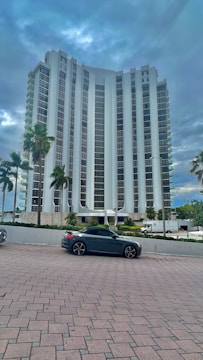 A tall, modern high-rise building with a sleek architectural design stands amidst a backdrop of cloudy skies. Several palm trees line the foreground near a parked dark-colored convertible car. The building is surrounded by a neat plaza with brick paving.