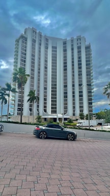 A tall, modern high-rise building with a sleek architectural design stands amidst a backdrop of cloudy skies. Several palm trees line the foreground near a parked dark-colored convertible car. The building is surrounded by a neat plaza with brick paving.