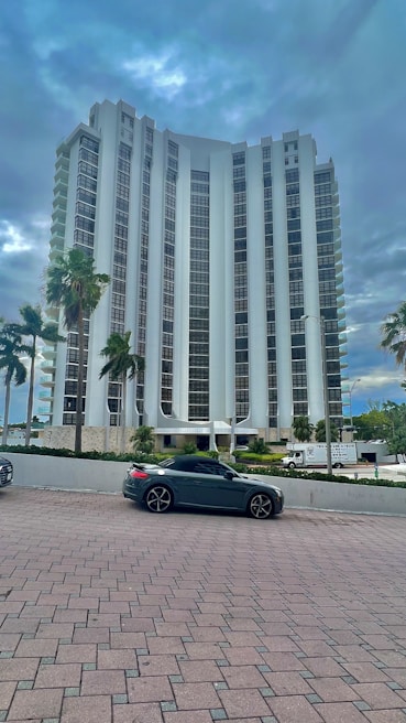 A tall, modern high-rise building with a sleek architectural design stands amidst a backdrop of cloudy skies. Several palm trees line the foreground near a parked dark-colored convertible car. The building is surrounded by a neat plaza with brick paving.