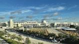 A large airport terminal with a white, modern architectural design set against a bright blue sky with scattered clouds. In the foreground, there is a parking lot full of yellow taxis and a wide road with vehicles, surrounded by greenery.