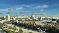 A large airport terminal with a white, modern architectural design set against a bright blue sky with scattered clouds. In the foreground, there is a parking lot full of yellow taxis and a wide road with vehicles, surrounded by greenery.