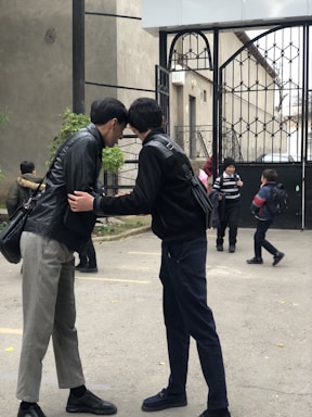 Two individuals are standing close together, appearing to have a private conversation. They are outdoors, near an iron gate and some greenery. In the background, a few children carry backpacks and walk, suggesting a school or public setting.