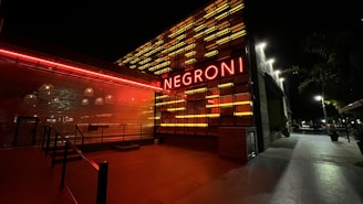 A modern building entrance with vibrant neon lights spelling 'Negroni'. The structure features a combination of red and yellow horizontal light strips, creating a striking visual against the dark night background. Streetlights and trees are visible in the distance along a sidewalk.