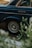A close-up view of the rear wheel of a vintage dark blue car, partially obscured by green foliage in the foreground. The visible car parts include a shiny silver hubcap displaying a logo and a chrome door handle. The scene is set on a street with subtle hints of pavement and road markings.