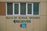 A beige wall with a row of four rectangular windows above a series of green text and symbols. The text includes a speed limit indication of 8 km/h, along with the words 'ON SCHOOL GROUNDS RECEIVER' and a left-pointing arrow. A blue sign with a white wheelchair symbol is also visible.