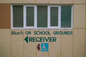 A beige wall with a row of four rectangular windows above a series of green text and symbols. The text includes a speed limit indication of 8 km/h, along with the words 'ON SCHOOL GROUNDS RECEIVER' and a left-pointing arrow. A blue sign with a white wheelchair symbol is also visible.
