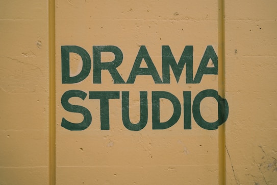 Bold green lettering spells out 'Drama Studio' against a muted yellow background, with visible texture and slight imperfections in the paint.