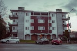 A mid-century style apartment building with a symmetrical facade. It has a combination of white walls and wooden paneling. There are balconies with metal railings on each side of the building. In front of the building, three parked cars are visible along the street. A lamppost and some city signs are seen on the sidewalk, and there are sparse trees and bushes around.