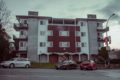 A mid-century style apartment building with a symmetrical facade. It has a combination of white walls and wooden paneling. There are balconies with metal railings on each side of the building. In front of the building, three parked cars are visible along the street. A lamppost and some city signs are seen on the sidewalk, and there are sparse trees and bushes around.