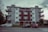A mid-century style apartment building with a symmetrical facade. It has a combination of white walls and wooden paneling. There are balconies with metal railings on each side of the building. In front of the building, three parked cars are visible along the street. A lamppost and some city signs are seen on the sidewalk, and there are sparse trees and bushes around.