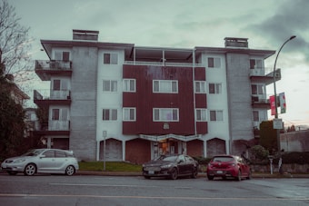 A mid-century style apartment building with a symmetrical facade. It has a combination of white walls and wooden paneling. There are balconies with metal railings on each side of the building. In front of the building, three parked cars are visible along the street. A lamppost and some city signs are seen on the sidewalk, and there are sparse trees and bushes around.