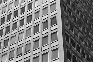 A high-contrast black and white photograph capturing the facade of a modern high-rise building with numerous square windows arranged in a geometric pattern. The angle of the shot emphasizes the repetition and symmetry of the windows, creating a sense of depth and uniformity.