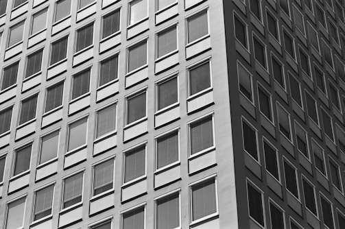 A high-contrast black and white photograph capturing the facade of a modern high-rise building with numerous square windows arranged in a geometric pattern. The angle of the shot emphasizes the repetition and symmetry of the windows, creating a sense of depth and uniformity.