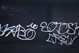 White graffiti text is scribbled across a dark surface. The text appears stylized, with flowing lines and loops creating an artistic effect. The background is dark, which allows the graffiti to stand out prominently.