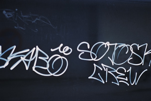 White graffiti text is scribbled across a dark surface. The text appears stylized, with flowing lines and loops creating an artistic effect. The background is dark, which allows the graffiti to stand out prominently.