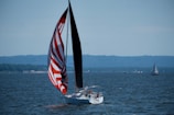 A sailboat with a striking red and white striped spinnaker glides across a vast body of water. The scene captures a calm day with other sailboats in the distance and a rugged, tree-lined coastline under a clear blue sky.