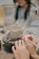 Hands are shaping a clay mug on a small rotating platform, which has two handles. A sculpting tool is being used to add details to the mug. In the background, a person appears to be observing or participating in the activity.