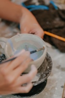 A person is carefully painting a ceramic cup using a fine brush. The cup is placed on a rotating platform, suggesting the process of crafting pottery. The hands appear focused, and there is a detailed design being applied inside the cup. Nearby materials like paint and fabric lie scattered around.