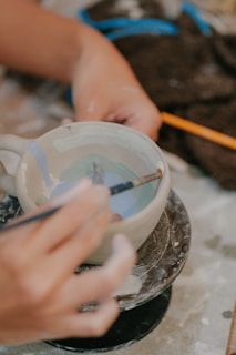 A person is carefully painting a ceramic cup using a fine brush. The cup is placed on a rotating platform, suggesting the process of crafting pottery. The hands appear focused, and there is a detailed design being applied inside the cup. Nearby materials like paint and fabric lie scattered around.