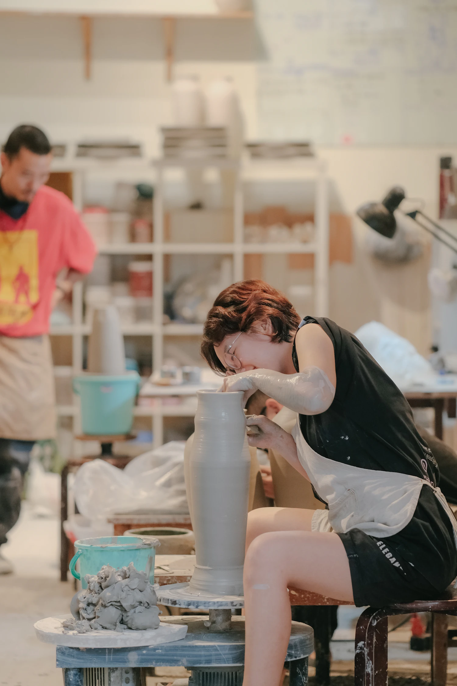 A woman throwing a tall clay vessel on a wheel in a bright studio, finished pieces drying on shelves behind