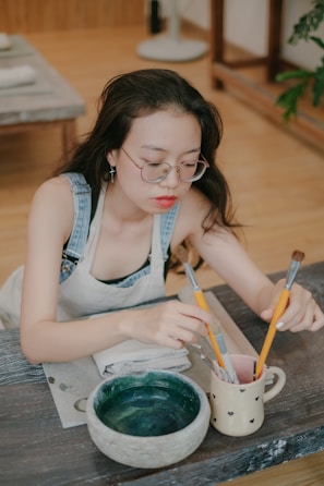 A young woman with glasses and long hair is seated at a table, wearing a denim apron. She is engaged in painting, holding several paintbrushes in a mug. On the table, there is a small pottery bowl with a greenish hue and a folded cloth.