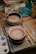 A ceramic bowl is on a pottery wheel with a spiral pattern in yellow and red. Surrounding the bowl are various pottery tools, brushes, and a palette with paint. A bowl filled with water and a brush is visible nearby. The scene takes place on a wooden table, suggesting a creative and artistic workspace.