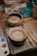 A ceramic bowl is on a pottery wheel with a spiral pattern in yellow and red. Surrounding the bowl are various pottery tools, brushes, and a palette with paint. A bowl filled with water and a brush is visible nearby. The scene takes place on a wooden table, suggesting a creative and artistic workspace.